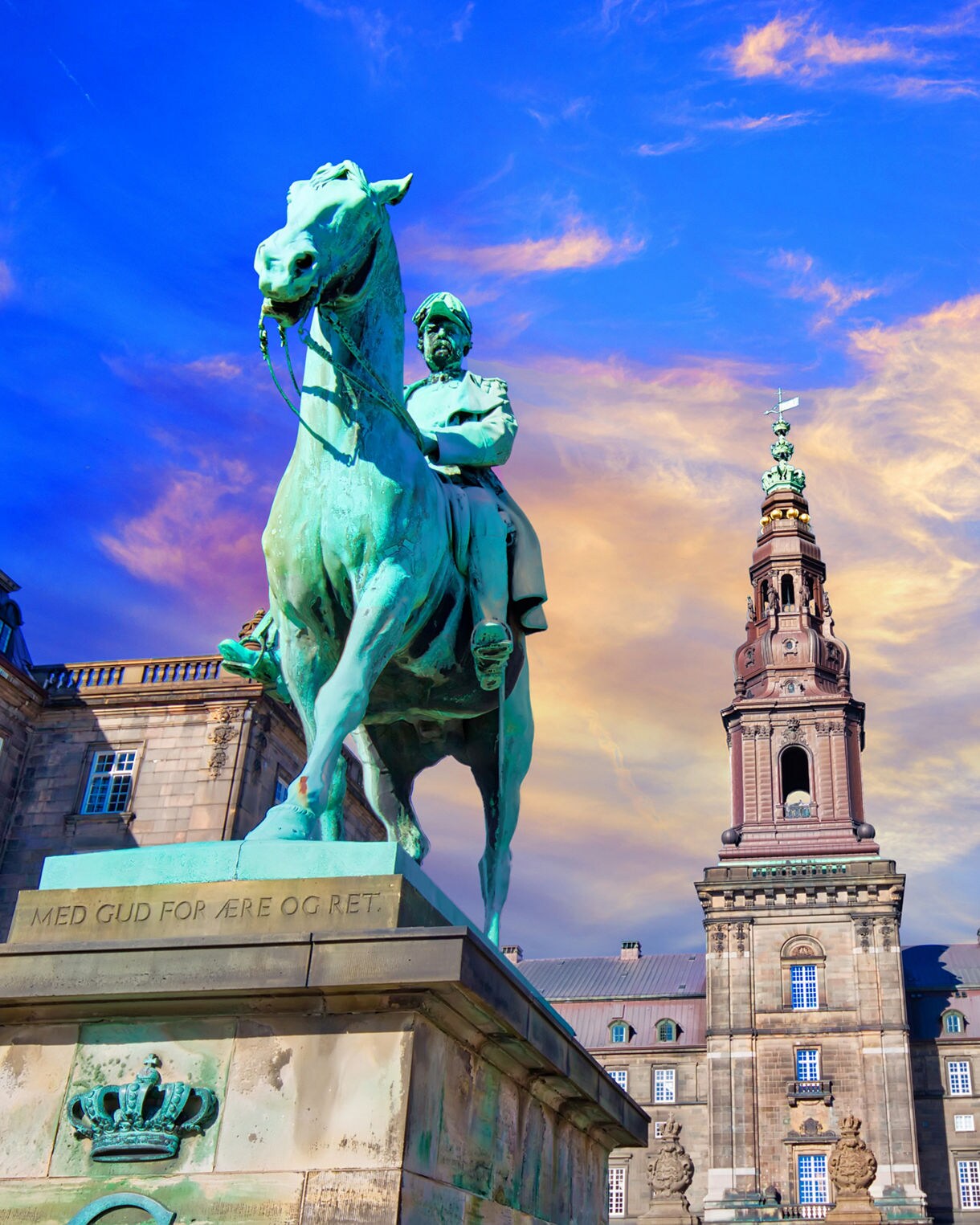 An equestrian statue of King Christian IX in front of Christiansborg Palace in Copenhagen, Denmark, with the palace tower rising in the background against a vibrant sunset sky.