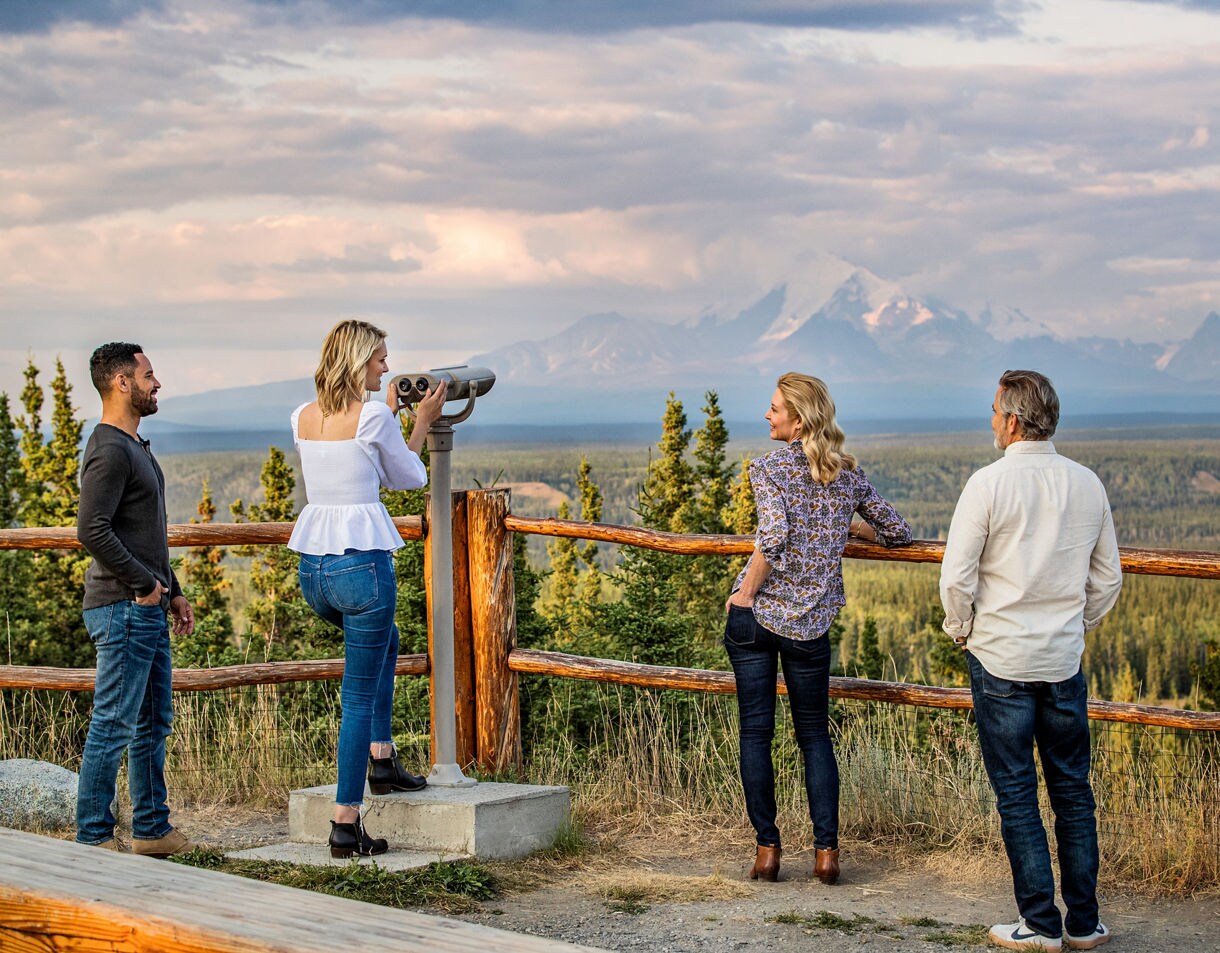 Two women at a mountain viewpoint, one looking through a coin-operated viewing scope while the other stands by a wooden fence railing, with snow-capped mountains and forests visible in the distance under a cloudy sky.
