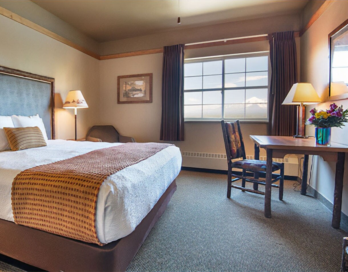 lean hotel room with a king bed featuring white linens and brown accent pillows, a small desk and chairs by the window, table lamps, and neutral-toned decor.