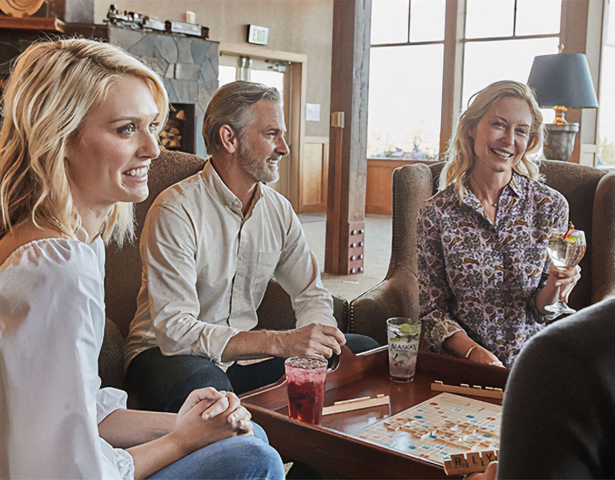 Three smiling guests sitting on comfortable chairs around a coffee table playing a board game with drinks, in a bright lodge interior with large windows and warm lighting.