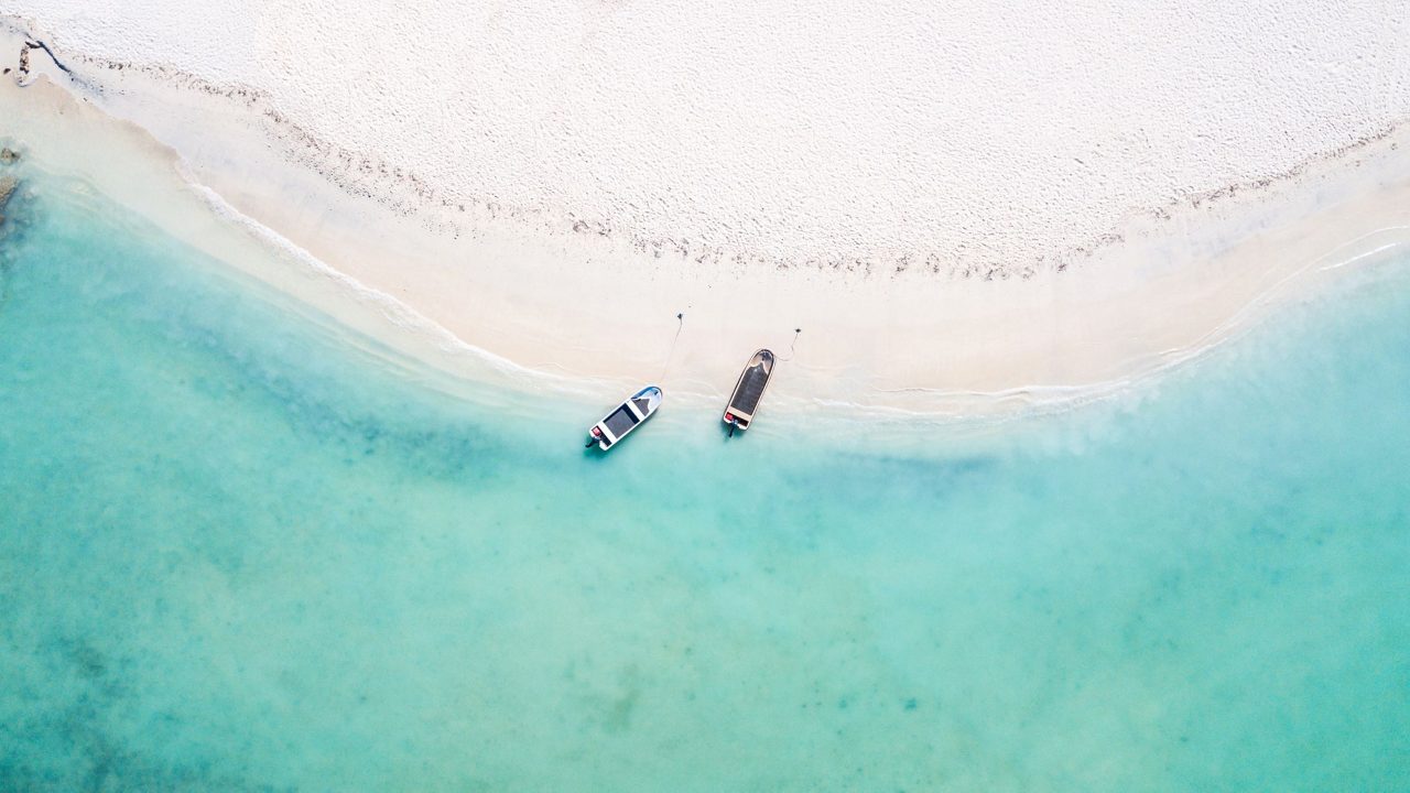 Aerial drone photograph showing two small boats floating in clear turquoise water near a curved white sand beach shoreline.