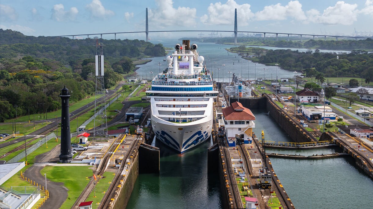 An aerial view of a Princess cruise ship passing through the Panama Canal locks, surrounded by green jungle hills and canal infrastructure.
