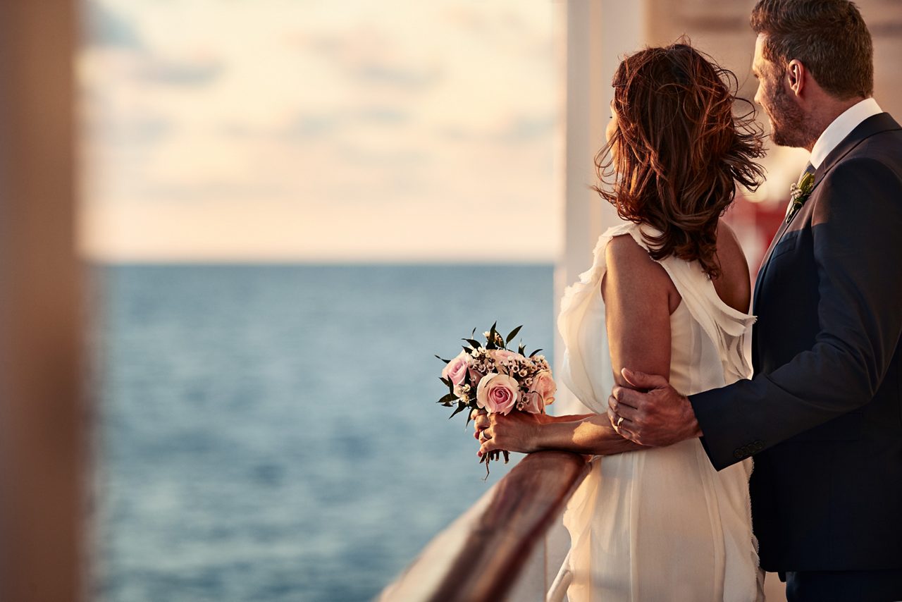 A bride and groom embrace on a balcony overlooking the ocean, the bride holding a small bouquet.
