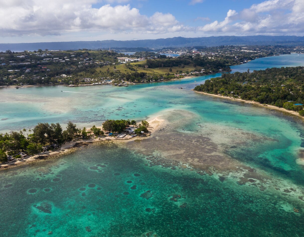 Aerial view of a shallow turquoise lagoon with coral patches, sandbars and a thin strip of land lined with trees, backed by rolling hills and scattered homes.