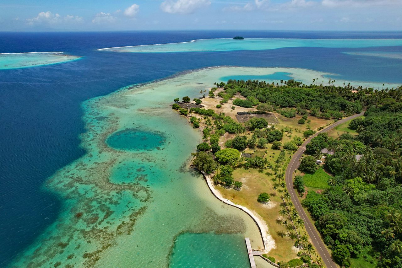 Aerial view of Raiatea’s coastline showing coral reefs, turquoise shallows and lush green landscape bordered by a winding road.