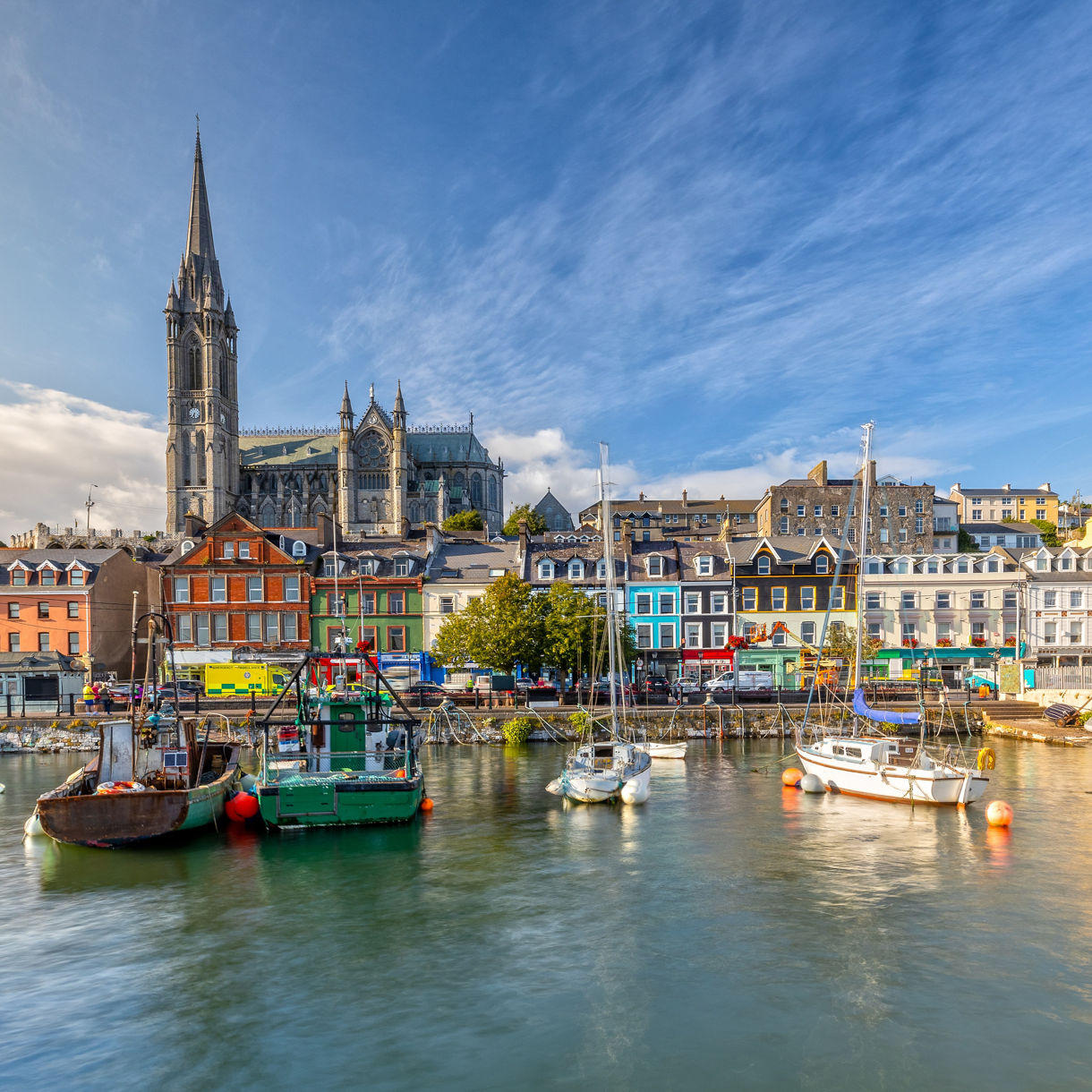 Waterfront view of Cobh, Ireland, with colorful row houses, boats moored in the harbor and St. Colman's Cathedral rising above the town.