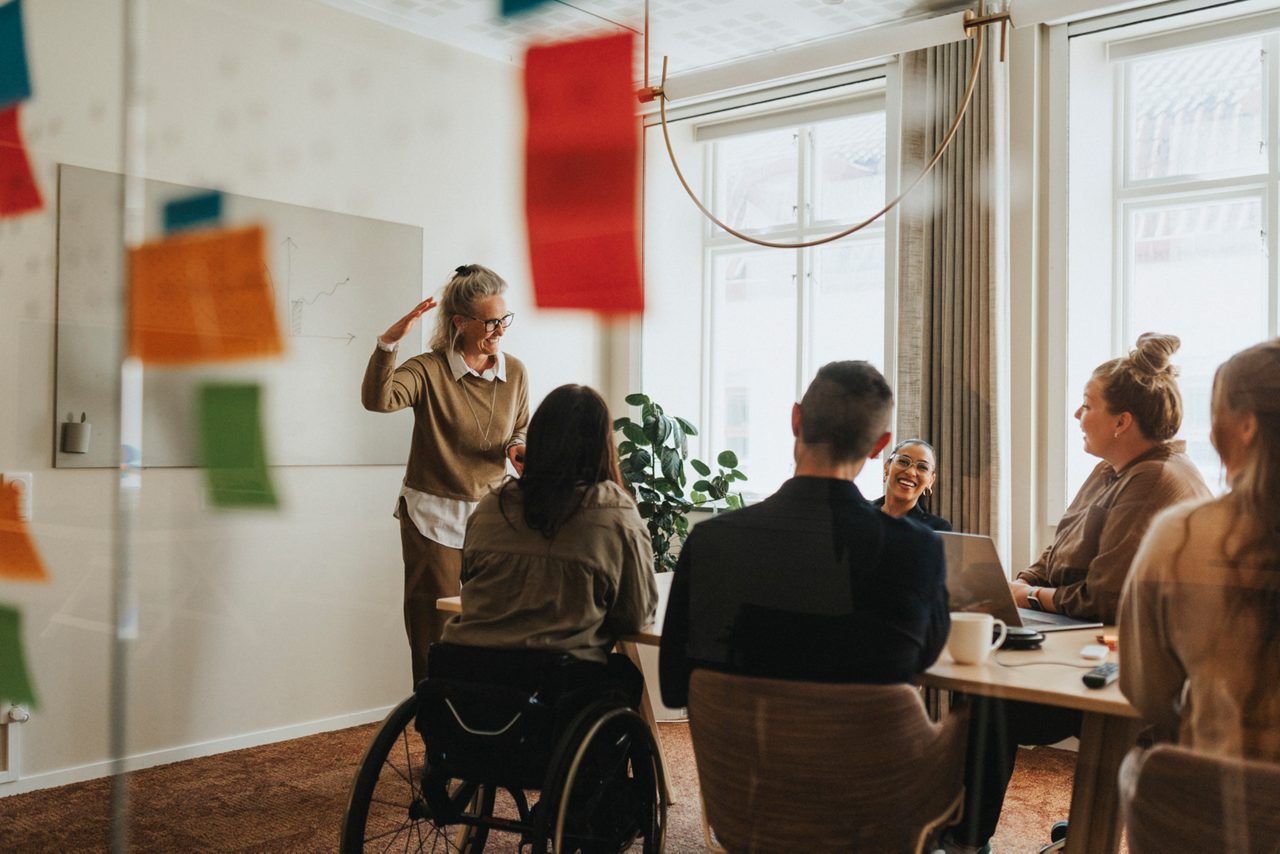 A diverse group of colleagues collaborating in a bright meeting room, with one participant using a wheelchair.