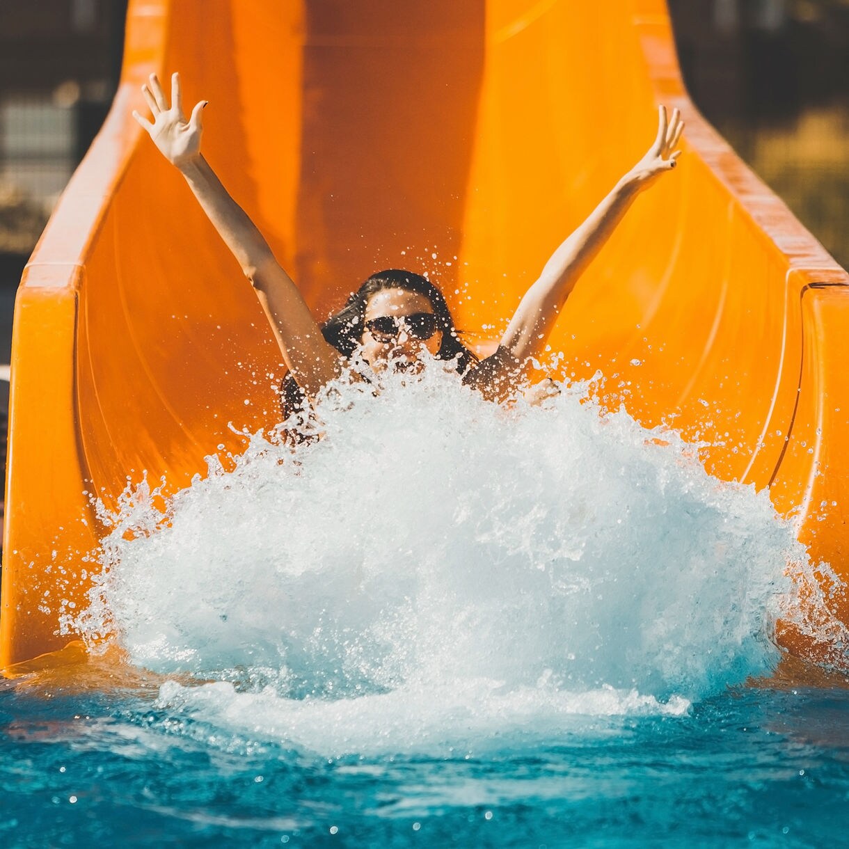 Woman with arms raised splashing into a pool at the bottom of a bright orange water slide at an outdoor adventure park in Costa Maya