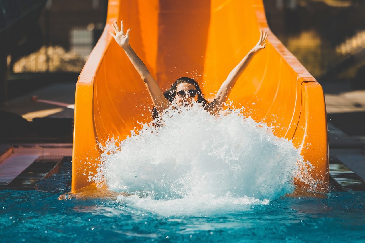 Woman with arms raised splashing into a pool at the bottom of a bright orange water slide at an outdoor adventure park in Costa Maya