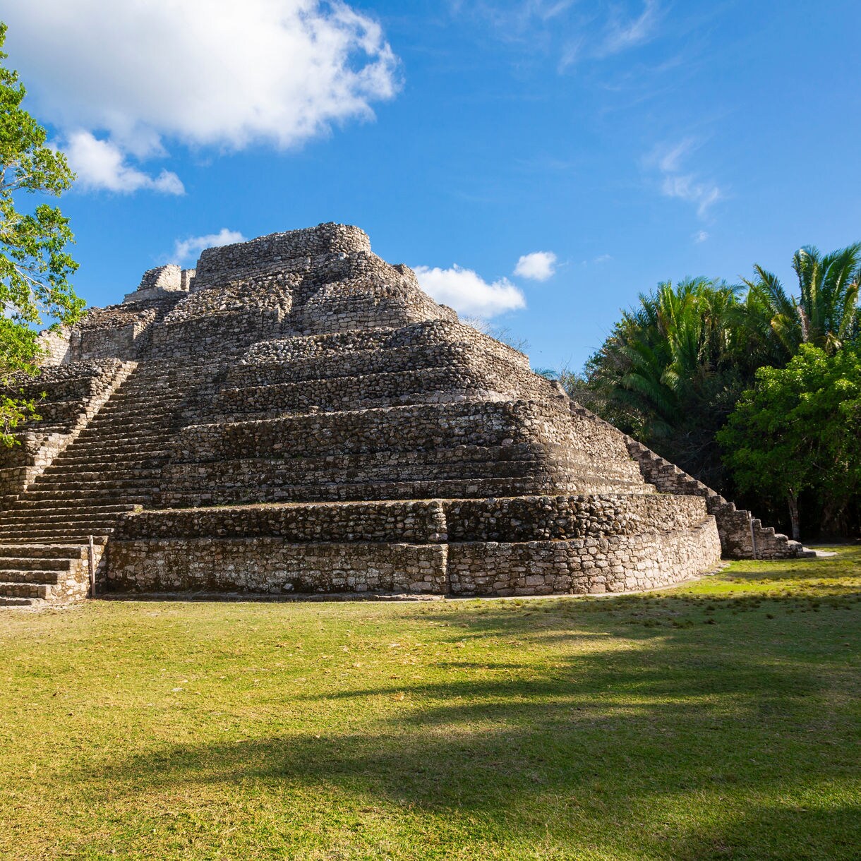 Large stepped pyramid structure made of stone at the Chacchoben Mayan ruins in Costa Maya, surrounded by grass and lush trees under a blue sky