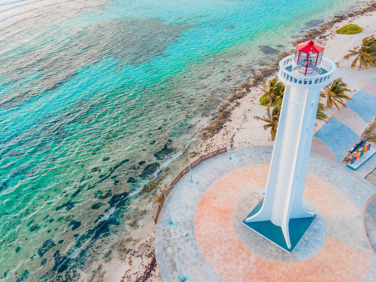 Aerial view of the white Faro de Mahahual lighthouse with a red top, standing beside a shoreline of clear blue water, coral reef, and sandy beach in Costa Maya