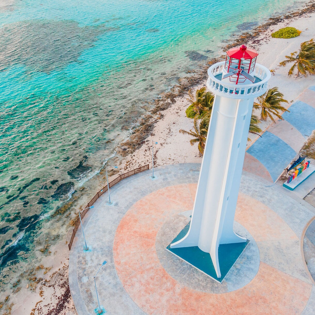 Aerial view of white lighthouse with red roof standing on sandy beach next to turquoise Caribbean waters.