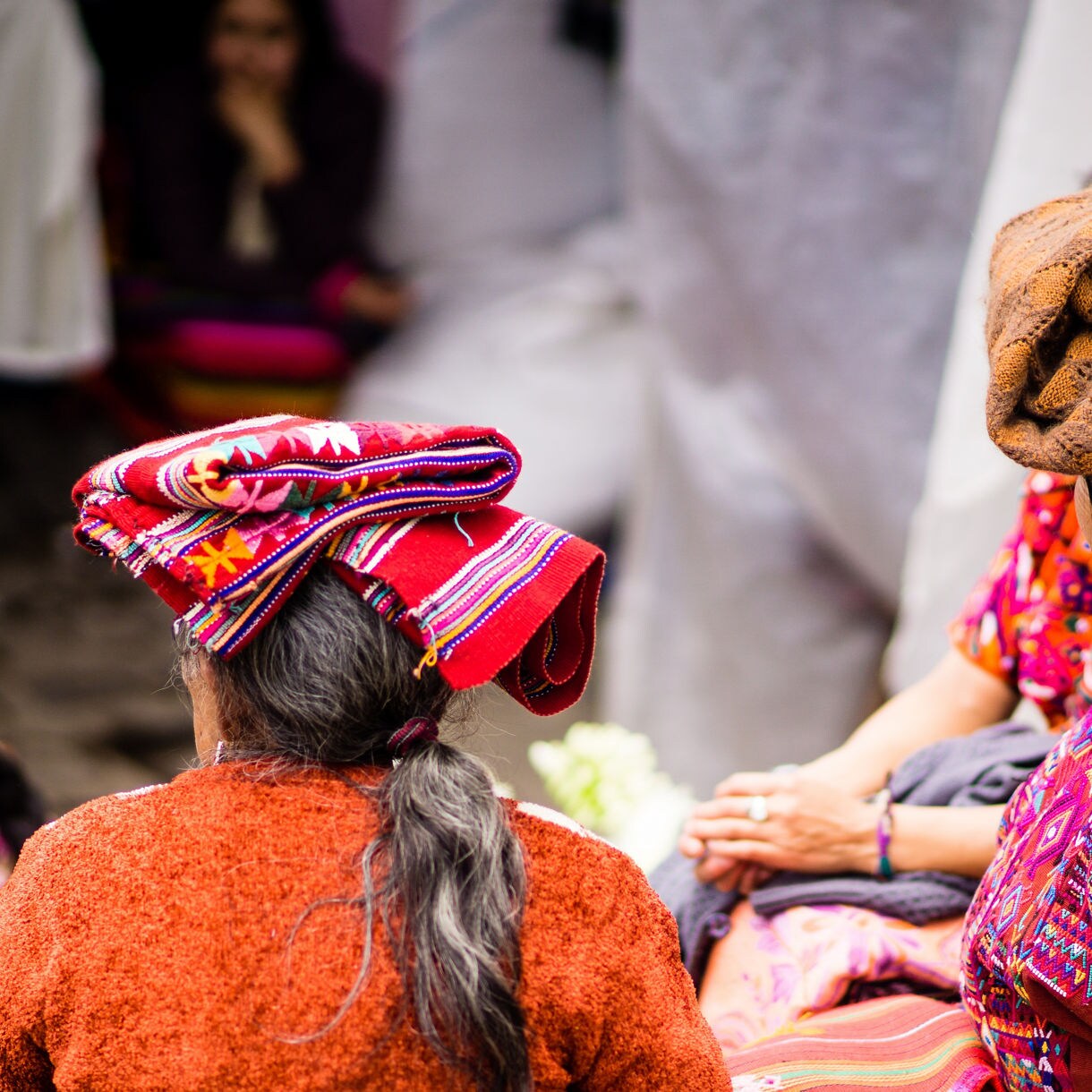 Close-up of two women wearing traditional Mayan clothing and woven headwraps, seated in a marketplace surrounded by colorful fabrics in Costa Maya