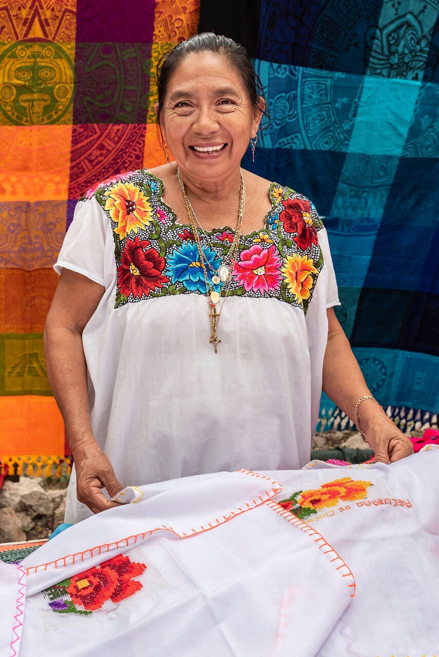Close-up of two women wearing traditional Mayan clothing and woven headwraps, seated in a marketplace surrounded by colorful fabrics in Costa Maya