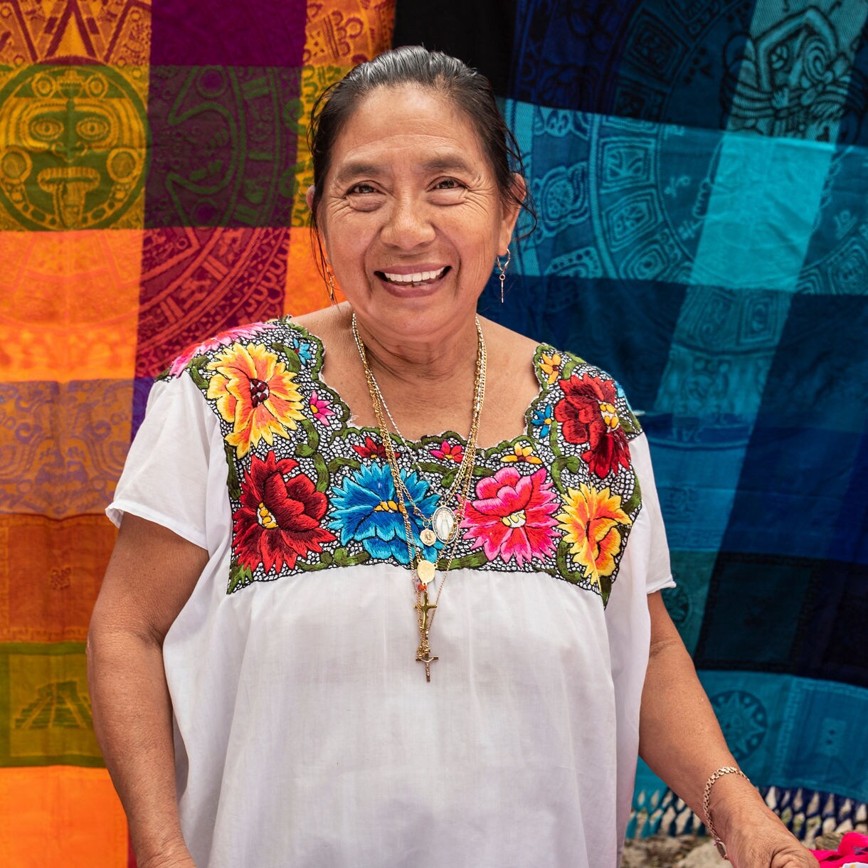 Smiling local woman wearing traditional colorful embroidered Mexican dress with floral patterns and gold jewelry.