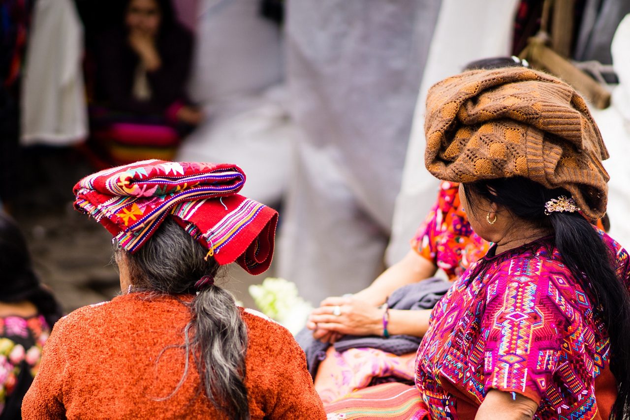 Close-up of two women wearing traditional Mayan clothing and woven headwraps, seated in a marketplace surrounded by colorful fabrics in Costa Maya