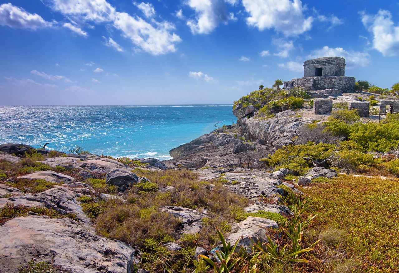 A stunning coastal view at Costa Maya, Mexico, showcasing ancient Mayan ruins perched on rocky cliffs by the sparkling blue ocean, surrounded by rugged terrain and lush greenery.