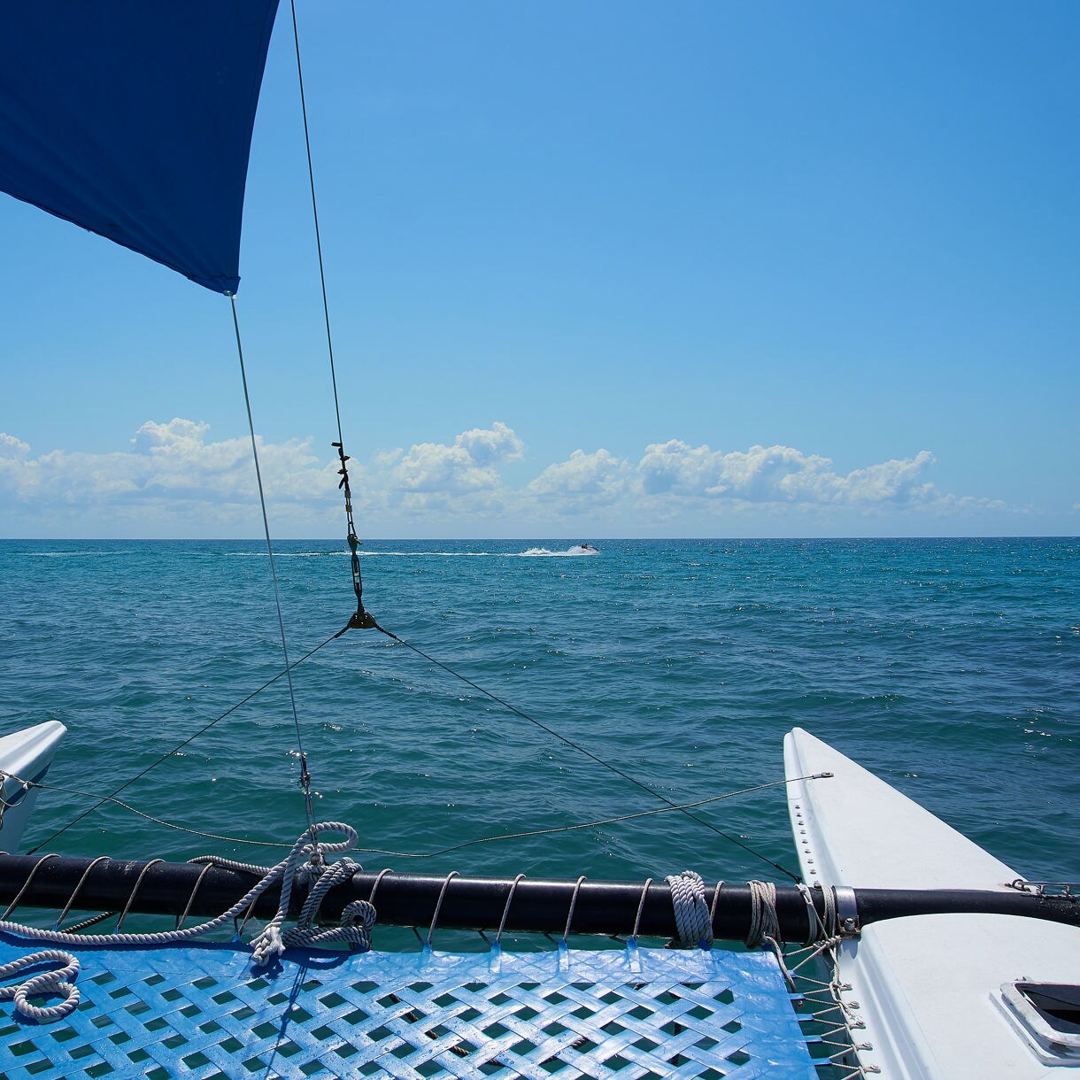 View from the front of a catamaran sailing on calm blue water with a blue mesh deck, ropes, and a sail against a backdrop of open sea and scattered clouds