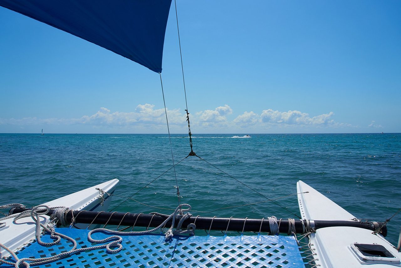 View from the front of a catamaran sailing on calm blue water with a blue mesh deck, ropes, and a sail against a backdrop of open sea and scattered clouds