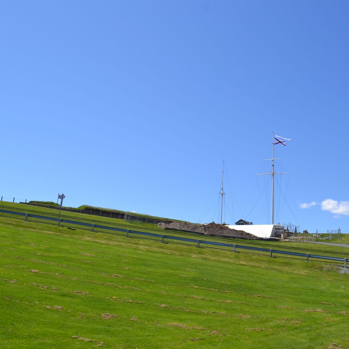 Grassy hill with fencing leading up to tall flagpoles and a Nova Scotia flag under a bright blue sky.