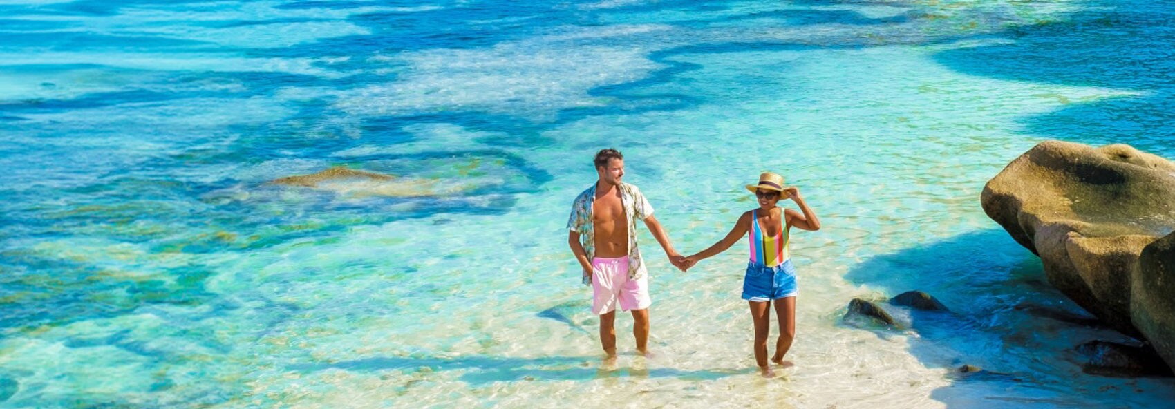 A joyful couple walking along a beach at the edge of clear turquoise water. They appear to be laughing and enjoying their time together in a tropical setting. The beach scene is calm, with the horizon visible under a cloudy sky.