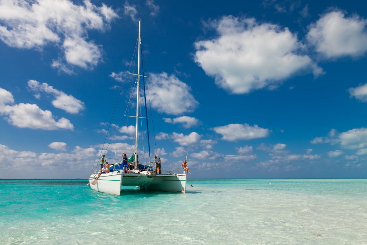 A group of people enjoy a catamaran ride on calm, turquoise waters off Cozumel, under a bright blue sky filled with fluffy clouds.