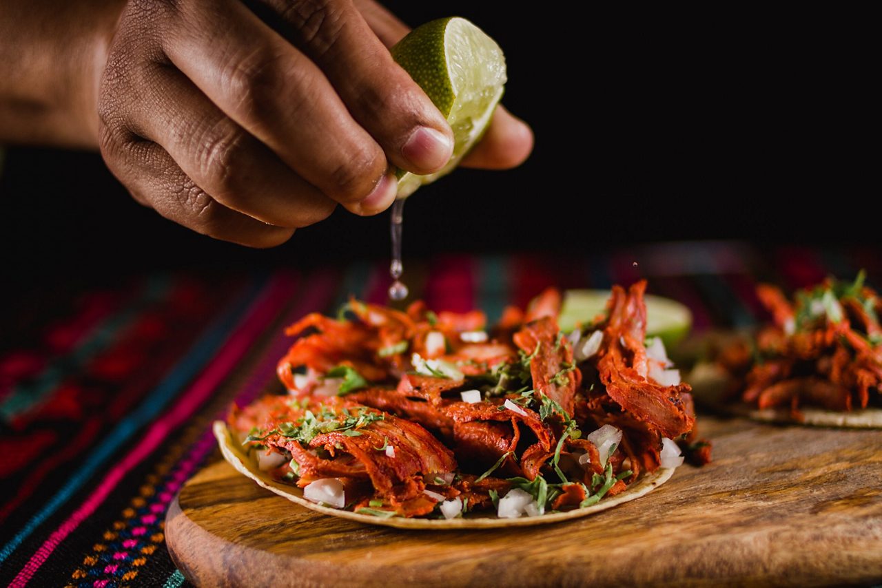 A close-up image of a hand squeezing lime onto a taco filled with seasoned shredded pork, cilantro, and onions, showcasing traditional Mexican street food served on a wooden plate.