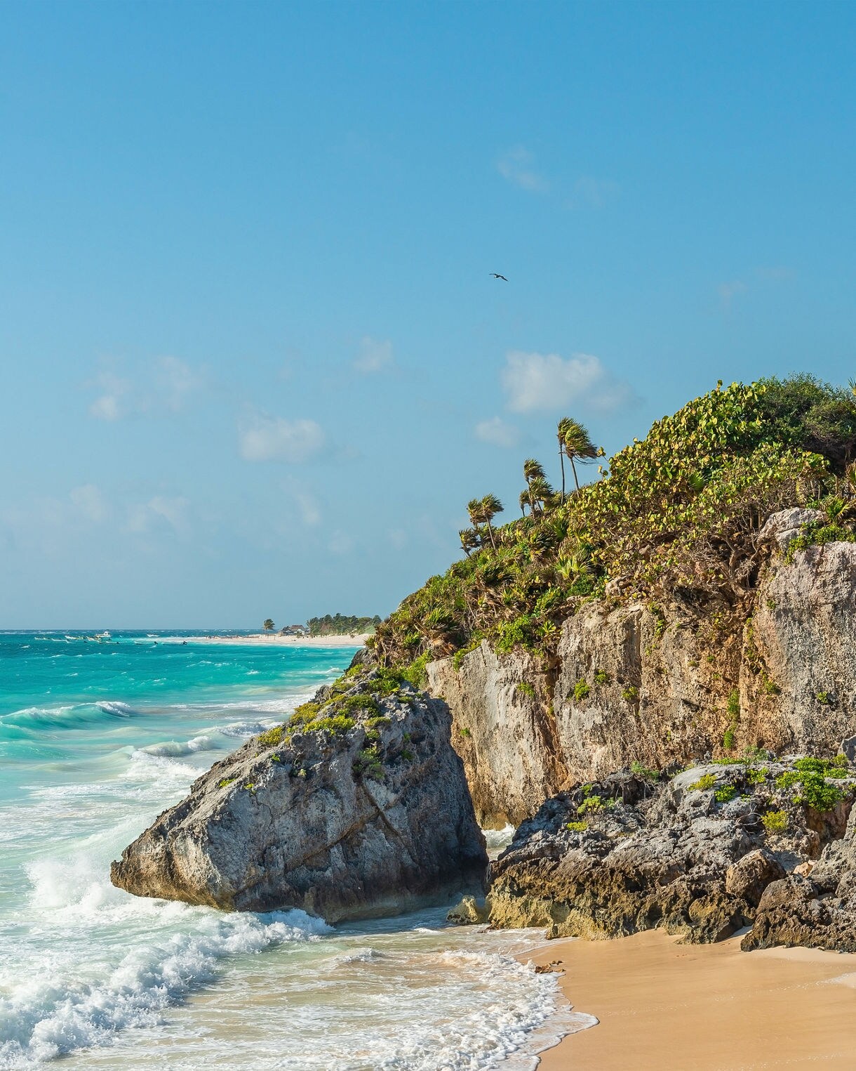 The Tulum Mayan ruins in Mexico sit atop a rocky cliff covered in lush greenery, overlooking a sandy beach and the turquoise waters of the Caribbean Sea