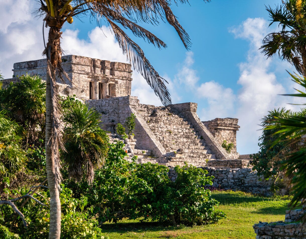 The Tulum Ruins in Cozumel, showcasing the ancient Mayan pyramid and steps, surrounded by lush greenery and palm trees against a bright blue sky