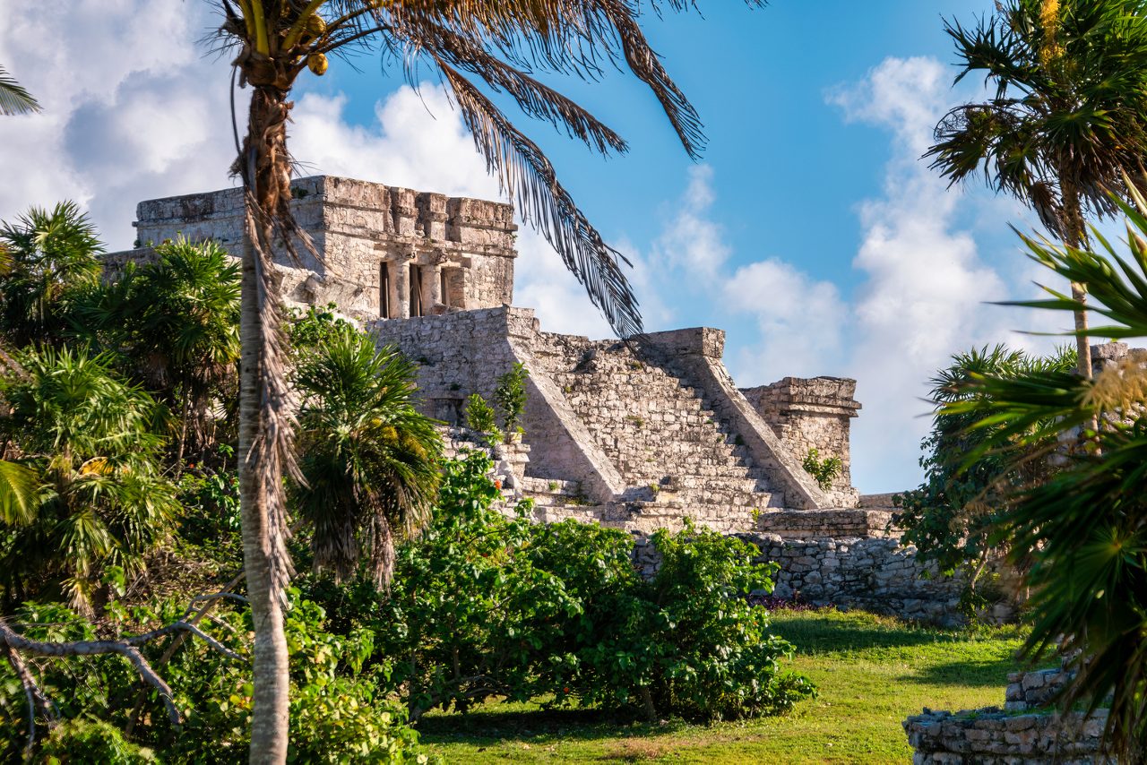 The Tulum Ruins in Cozumel, showcasing the ancient Mayan pyramid and steps, surrounded by lush greenery and palm trees against a bright blue sky