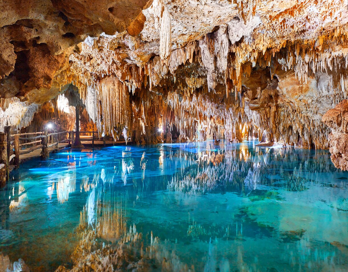 Stalactites hanging from the ceiling of a cave, reflected in the crystal-clear water below. Wooden walkways guide visitors through the scenic underground river at Xcaret Eco Archaeological Park in Cozumel.