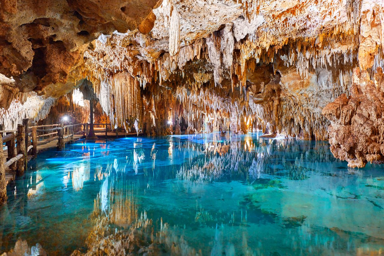 Stalactites hanging from the ceiling of a cave, reflected in the crystal-clear water below. Wooden walkways guide visitors through the scenic underground river at Xcaret Eco Archaeological Park in Cozumel.