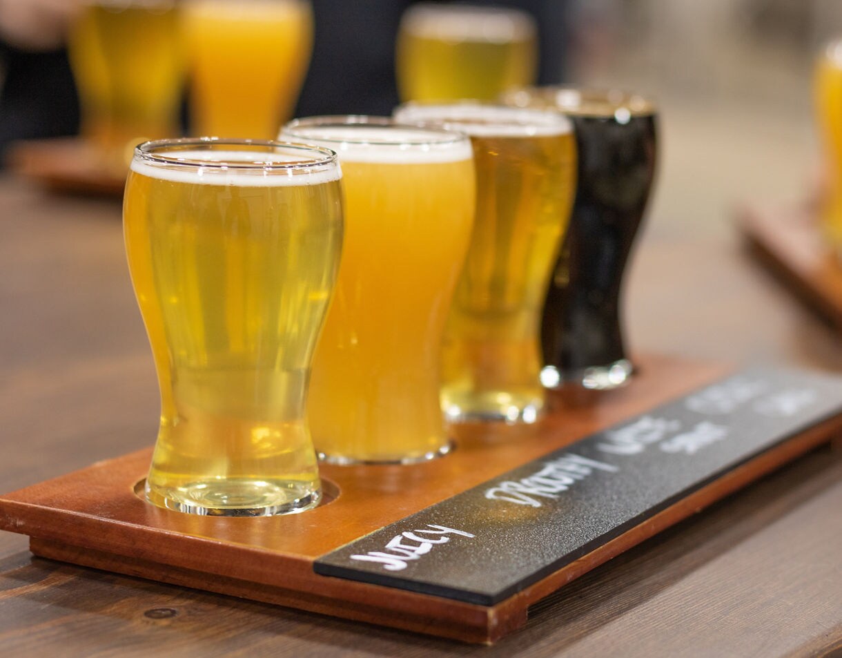 Close-up of a flight of craft beers in small glasses ranging from pale gold to dark amber on a wooden tasting tray.