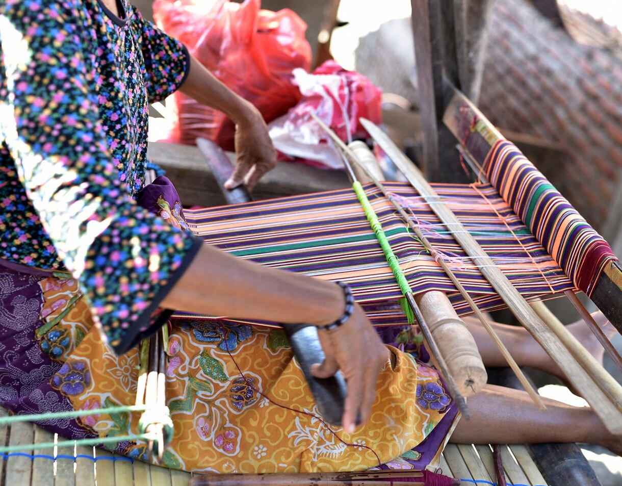 Close-up of a woman weaving colorful striped textile on a traditional loom, her hands guiding threads while she sits in patterned clothing.