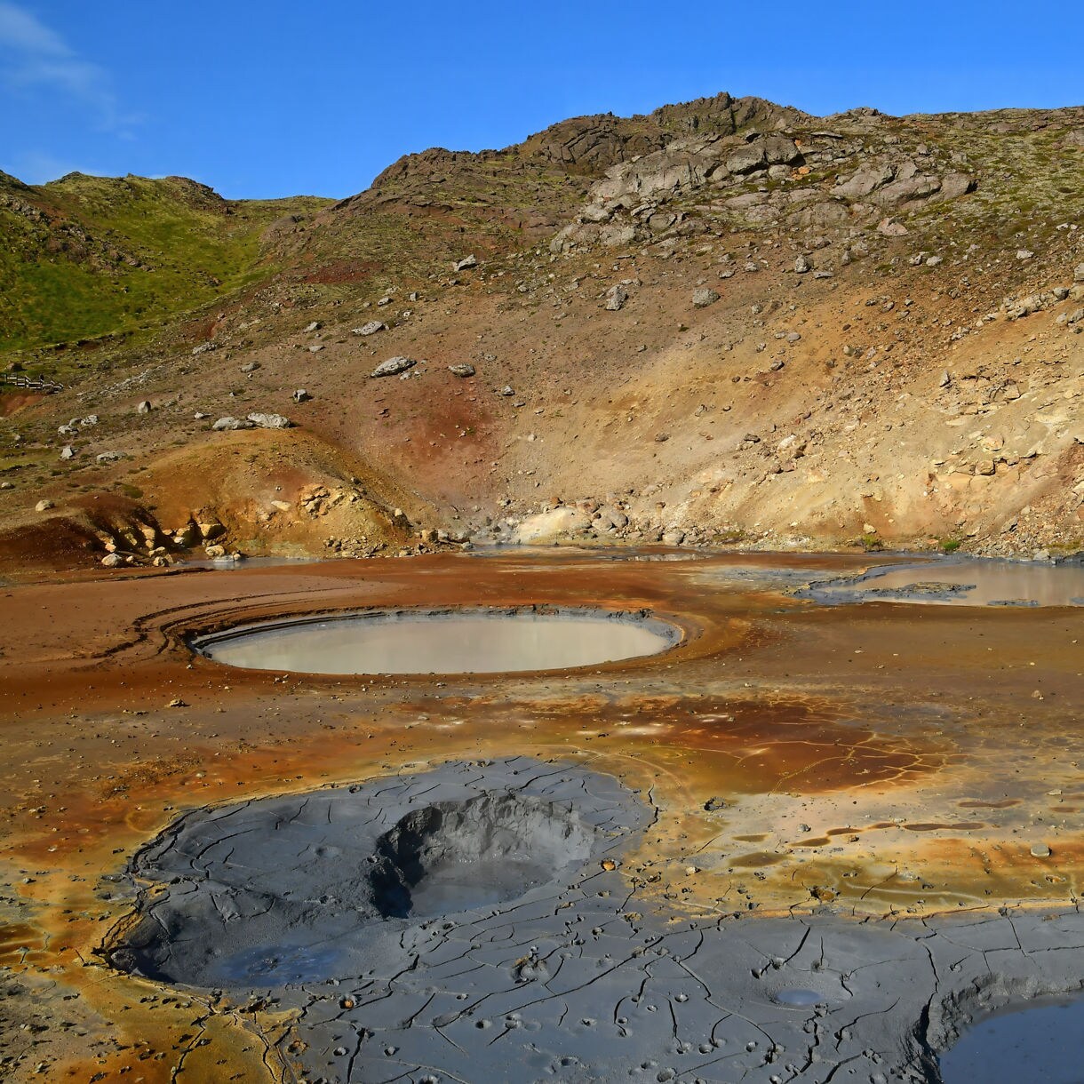 Geothermal area with steaming mud pools and colorful mineral deposits in a rocky Icelandic valley under a clear blue sky.