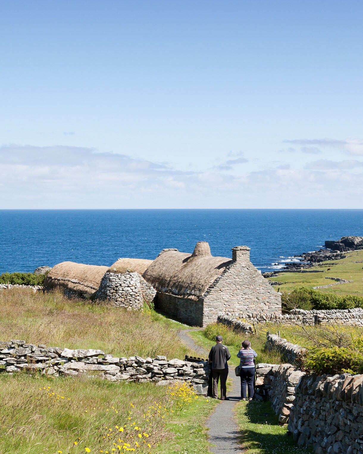 Stone croft houses with thatched roofs near the sea in Shetland, with two visitors walking down a narrow path.
