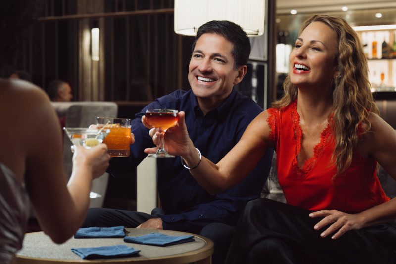 Two guests share a laugh while raising colorful cocktails in a modern lounge setting, with one person wearing a navy shirt and another in a red dress. 