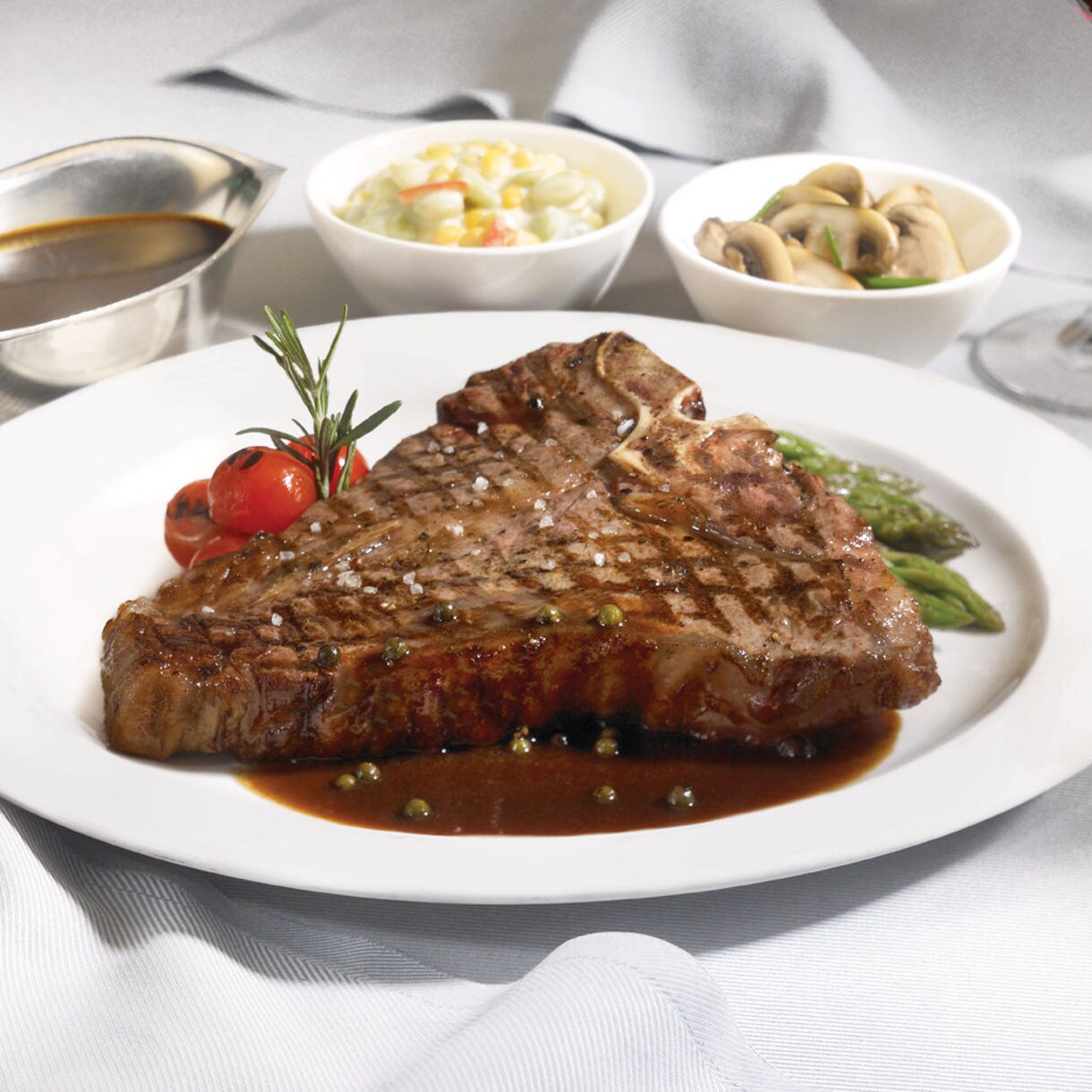 A perfectly grilled T-bone steak served on a white plate with a garnish of cherry tomatoes and asparagus. The meal is accompanied by side dishes including sautéed mushrooms, creamed corn, and a bowl of rich brown sauce. The setting features a fine dining ambiance with a glass of red wine in the background.