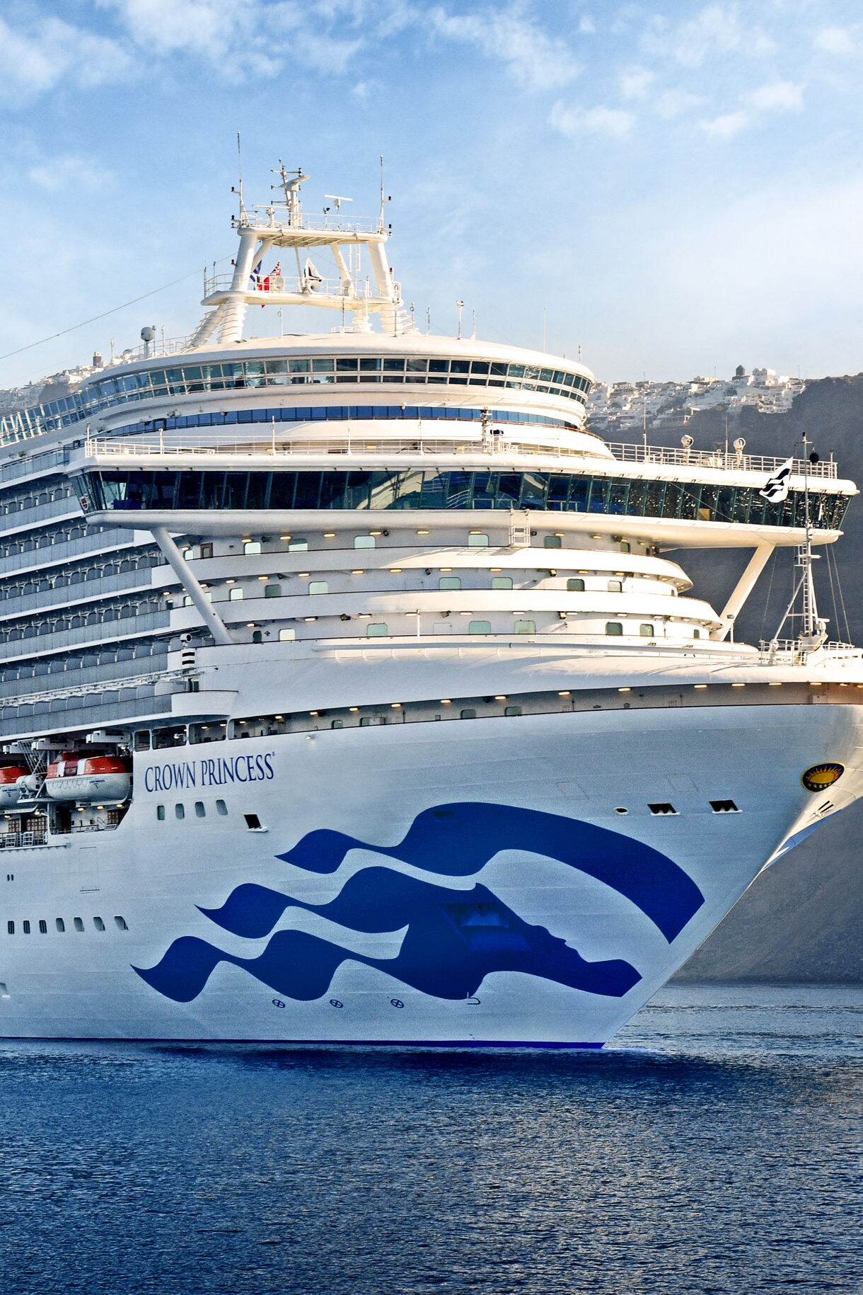 Close-up view of the Crown Princess cruise ship at sea, highlighting its iconic blue wave logo on the bow, with the coastline and white buildings in the background.