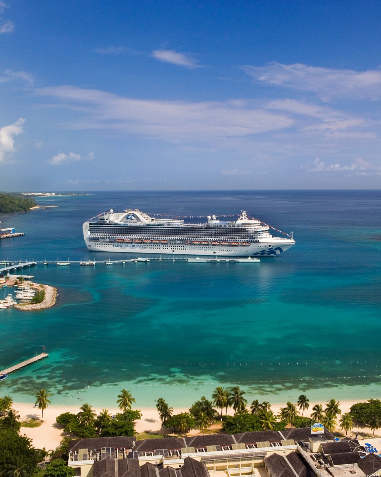 Aerial view of the Crown Princess cruise ship docked in the clear, turquoise waters of Ocho Rios, Jamaica, with white sandy beaches, palm trees, and lush green hills nearby.