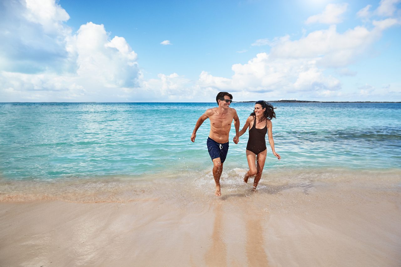 A joyful couple walking along a beach at the edge of clear turquoise water. They appear to be laughing and enjoying their time together in a tropical setting. The beach scene is calm, with the horizon visible under a cloudy sky.