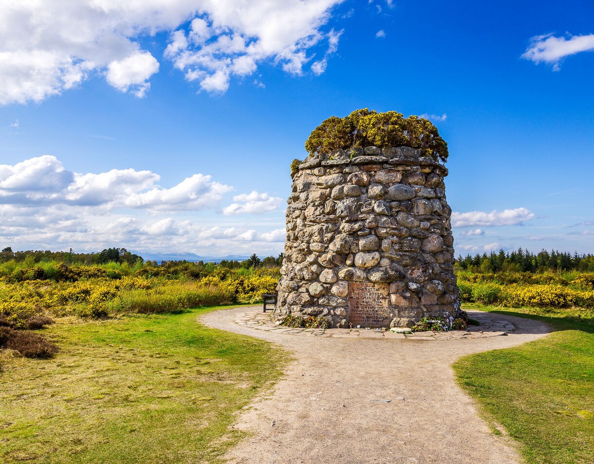 Stone cairn monument at Culloden Moor near Inverness, surrounded by grassy fields and wildflowers under a bright blue sky with scattered clouds.