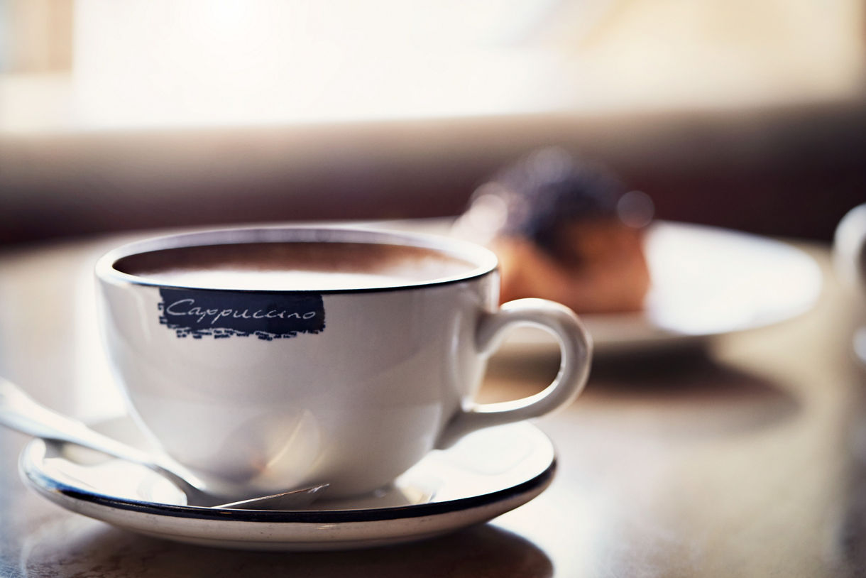 A cappuccino in a white branded cup and saucer on a wooden table with a blurred pastry in the background.