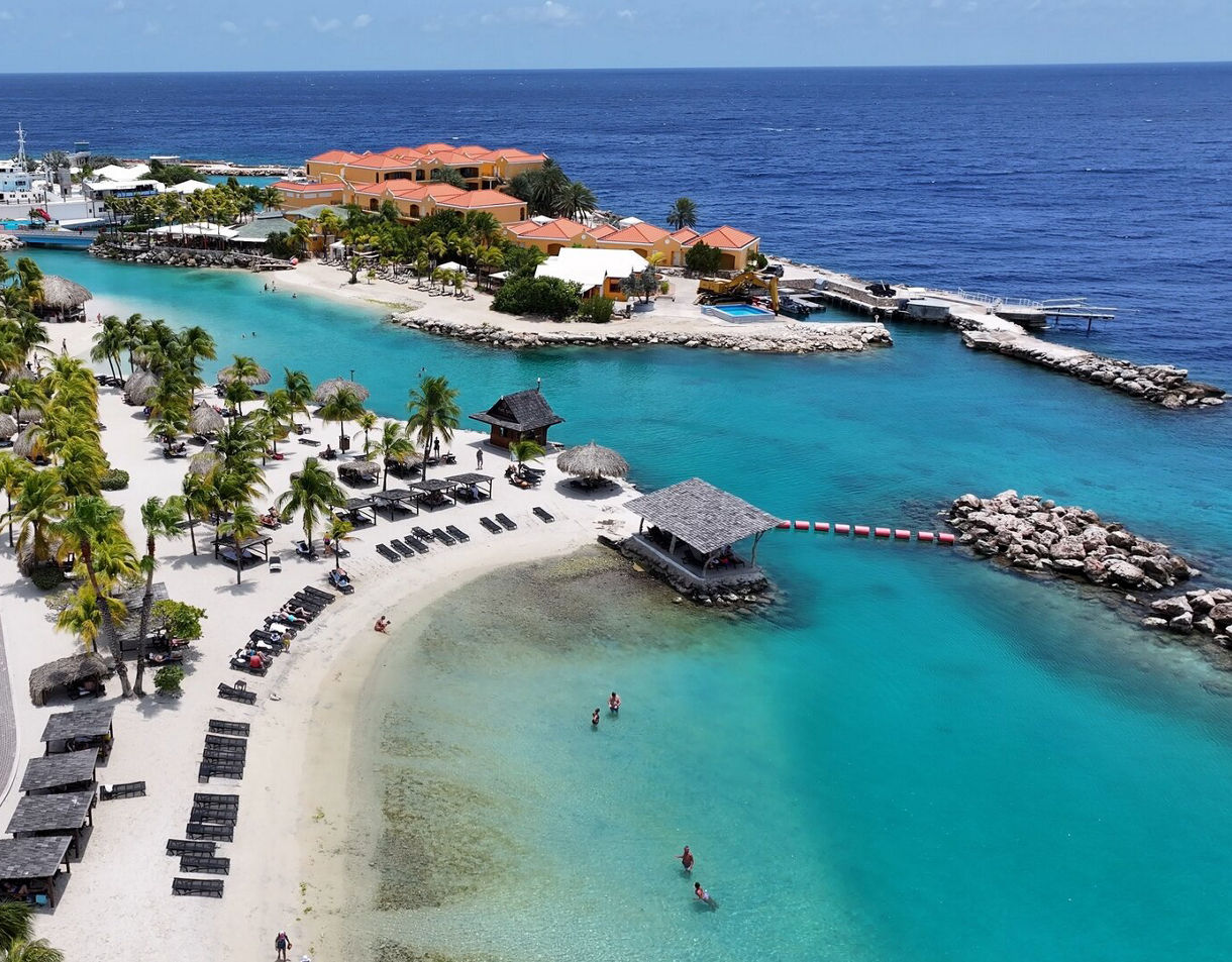 Aerial view of Blue Bay Beach in Curaçao featuring turquoise waters, white sandy shores, palm trees, shaded cabanas and a marina with boats in the background.