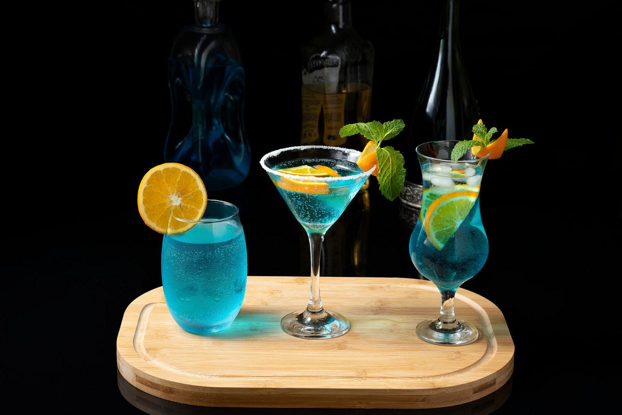 Three Blue Curaçao cocktails garnished with orange slices, lime and fresh mint, elegantly arranged on a wooden tray against a dark background with bottles in the distance.