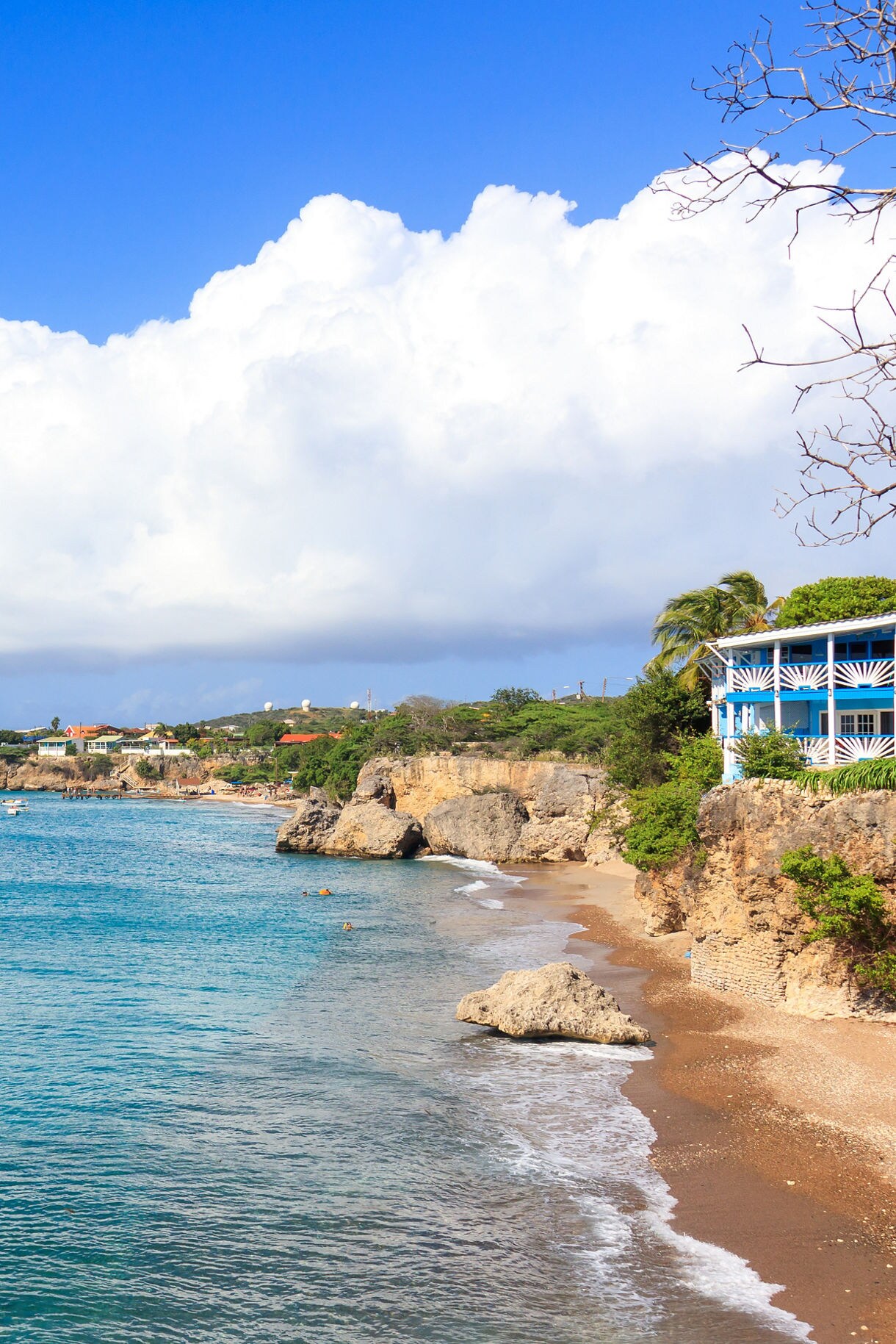 A picturesque view of Playa Kalki Beach in Curaçao, featuring calm turquoise waters, rocky cliffs, and a shoreline dotted with boats. A charming blue building overlooks the beach, with lush greenery and a bright sky in the background.