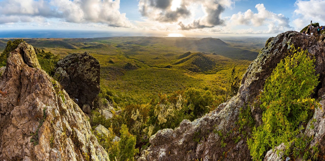A stunning view from the rocky peak of Christoffel National Park in Curaçao, overlooking rolling green hills, dense vegetation and the distant coastline under a golden sunset.