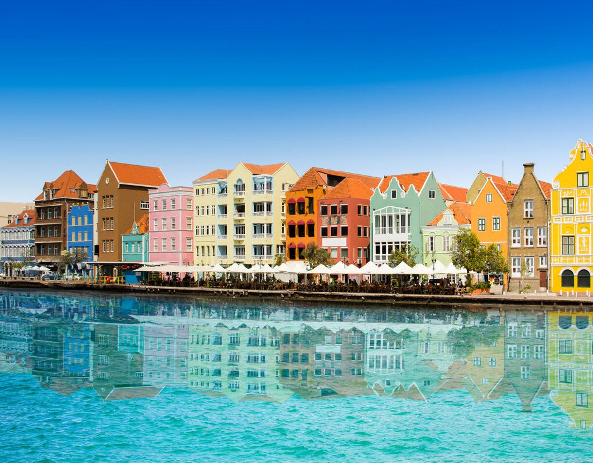 A row of brightly colored Dutch colonial buildings along the Handelskade waterfront in Willemstad, Curaçao, with their reflections shimmering in the clear blue water.