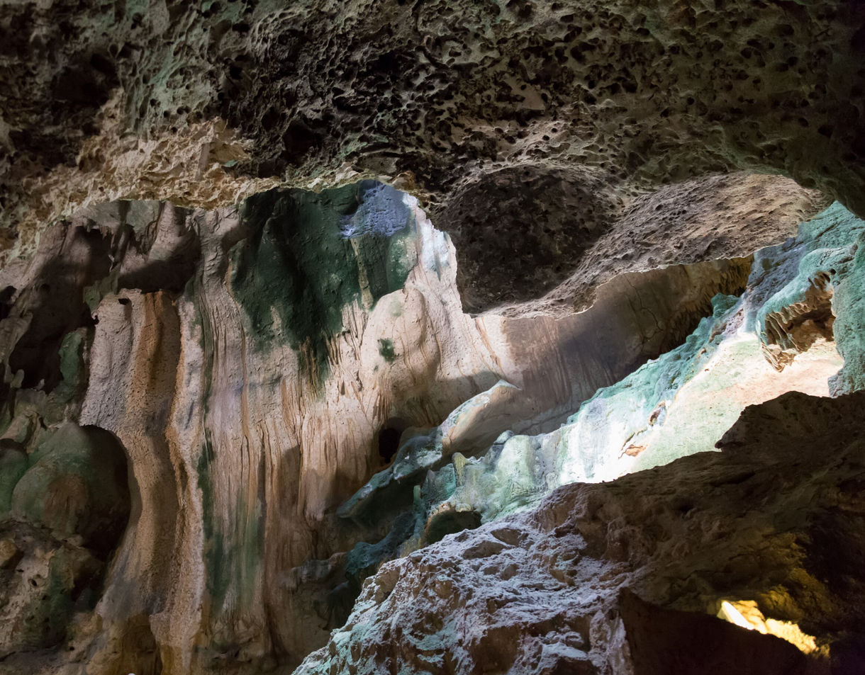 The interior of Hato Caves in Curaçao, showcasing rugged limestone formations with green moss, stalactites and natural light filtering through an opening in the cave ceiling.