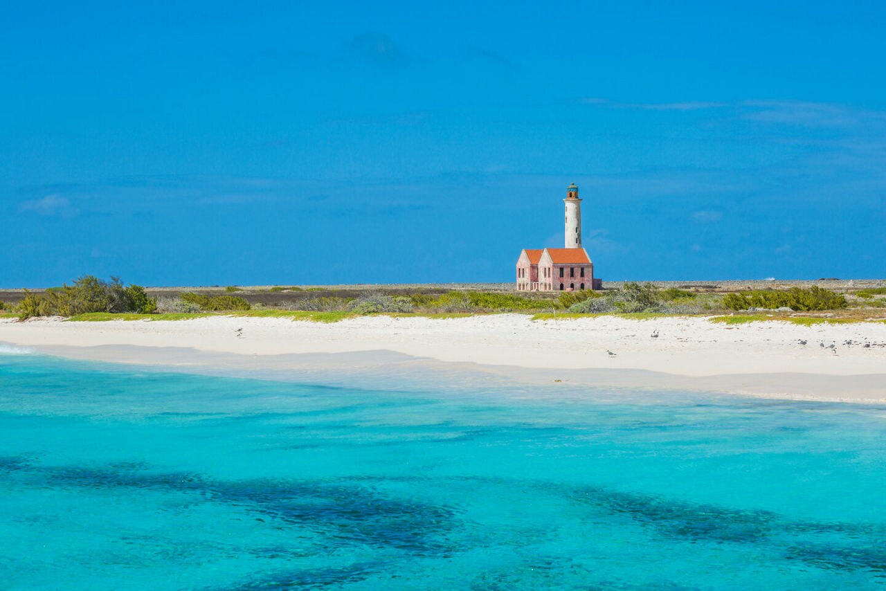 A remote beach on Klein Curaçao with crystal-clear turquoise waters, a stretch of white sand and an old weathered lighthouse with a red roof standing in the distance.